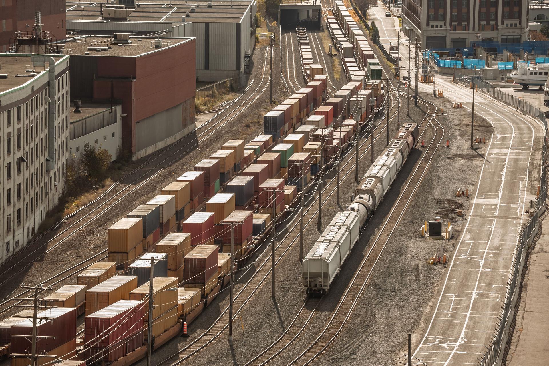 Container trains, hopper cars occupy multiple tracks in a railyard at the Port of Montreal, Quebec. Stacked boxes, and nearby warehouses illustrate North American logistics, exports and imports.